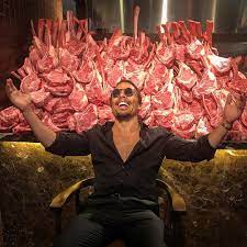 Nusret in an all black outfit and eyeglasses with meats behind him