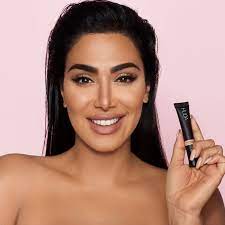 Huda Kattan in a pink background, smiling while holding a concealer